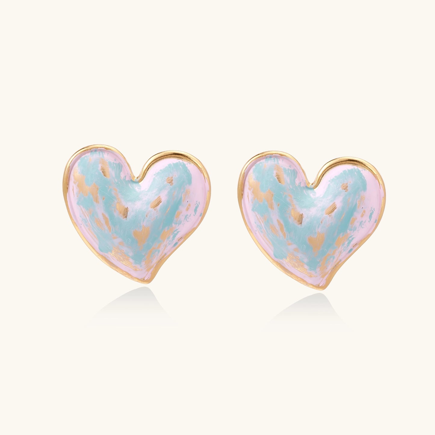Heart-shaped stud earrings with iridescent blue-pink enamel finish and gold-plated brass frames against cream background