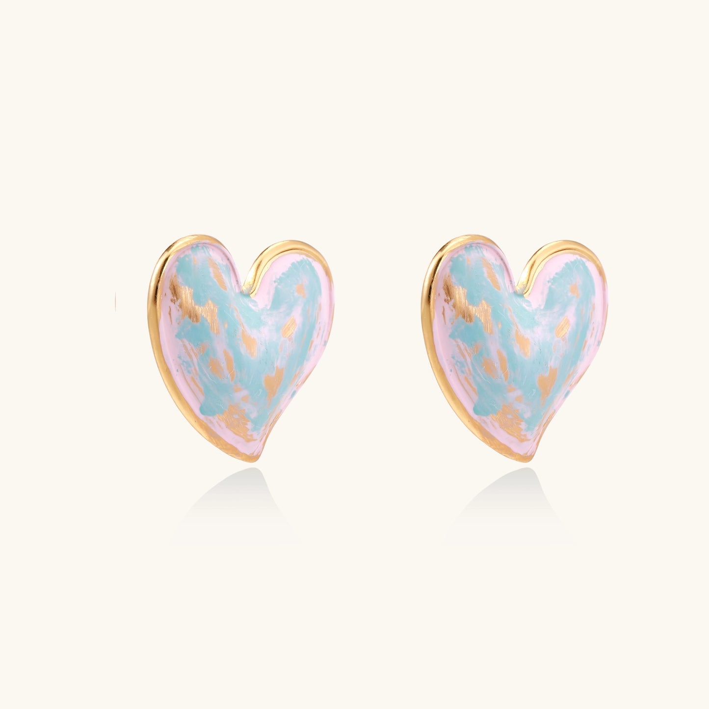 Heart-shaped stud earrings with iridescent blue-pink enamel finish and gold-plated brass frames against cream background