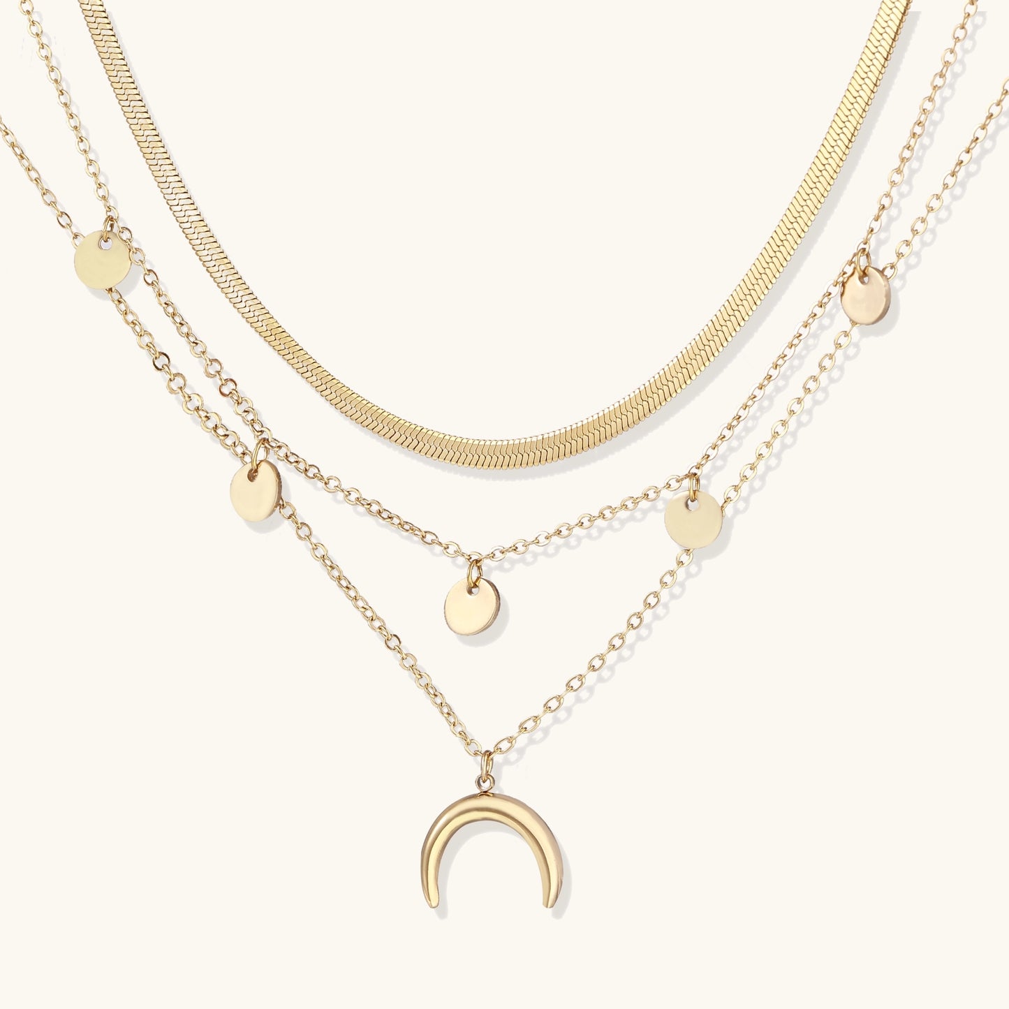 Gold-plated layered necklace set featuring herringbone chain, disc pendants, and crescent moon charm on beige background.