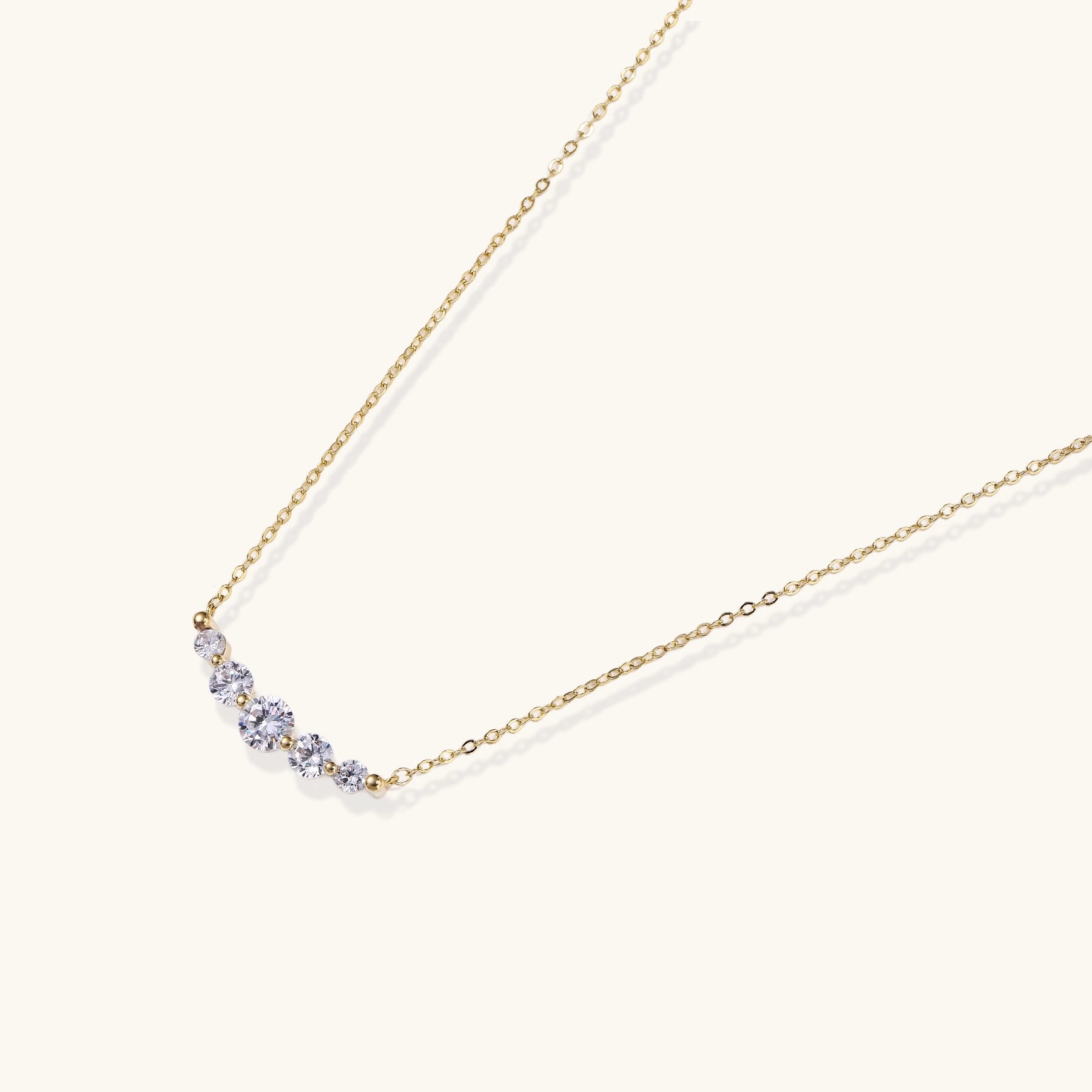 Delicate 14K gold plated necklace with curved arc of sparkling zirconia stones on cream background.